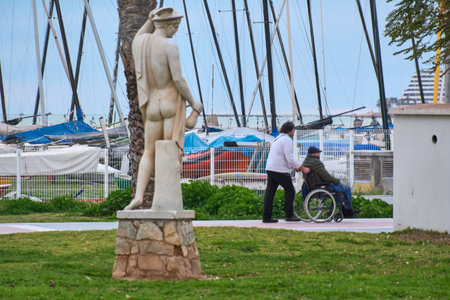 Sitges. Spain - February 17, 2026: Caregiver pushing elderly man in wheelchair along seaside promenade near marina with sailboats and classical statue in foreground.のeditorial素材
