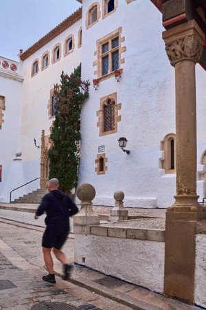 Sitges. spain - february 18, 2026: Runner passing historic whitewashed building in sitges old town, cobblestone street and mediterranean architectural detailsのeditorial素材