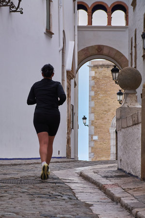 Sitges. spain - february 18, 2026: Rear view of woman training along narrow street in sitges historic center, urban fitness and coastal heritage settingのeditorial素材