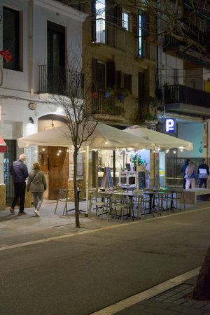 Sitges. Spain - March 13, 2026: Outdoor restaurant terrace with tables and umbrellas on a pedestrian street at night with people walking.のeditorial素材