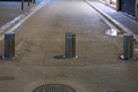 Three stainless steel automatic rising bollards installed on a stone paved street at night to...