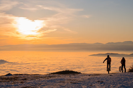 Two men on unicycles riding on snowy mountain Schoeckl in Styria, Austria over low stratus fog to...
