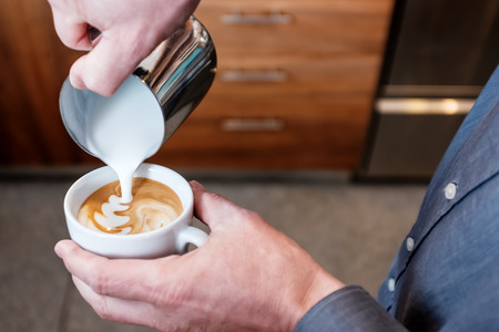 Professional barista pouring steamed milk from stainless steel tumbler into coffee cup making latte art on cappuccinoの写真素材