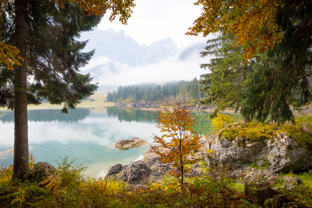 View over lake Laghi di Fusine on a foggy morning with some rocks at the beach and mountain range Mangart near Tarvisio in Italyの写真素材