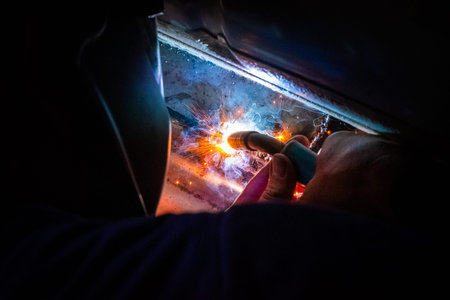 Man with a helmet and no gloves welds in the metal of a car and repairs a rusty hole