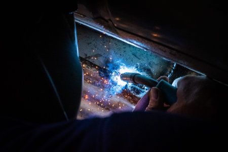 Man with a helmet and no gloves welds in the metal of a car and repairs a rusty hole