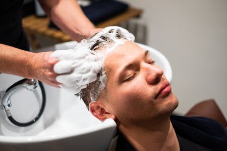 Barber washes young man's hair with lots of foam in his barbershop. Massages the head over the sink.の写真素材