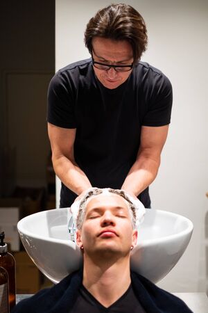 Barber washes young man's hair with lots of foam in his barbershop. Massages the head over the sink.の写真素材