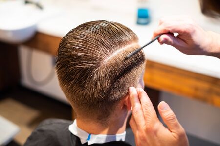 Barber is hairdressing the hair of a young man with a comb in his barbershop, ready to cut hairの写真素材