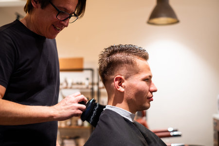 Male hairdresser brushes the hair of young man after cutting hair from the shoulder in his barbershopの写真素材