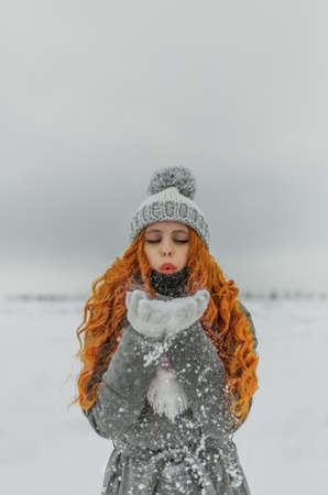 Ginger young beautiful woman is blowing snow from hands. Winter snowy day. Snowflakes are flyingの写真素材
