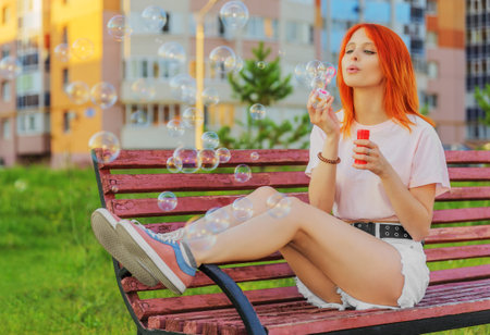 Beautiful young red-haired woman sitting on bench and making blowing bubblesの写真素材