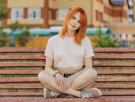 Beautiful young red-haired woman sitting on bench and looking at camera. Summer eveningの写真素材