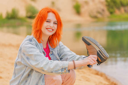 Beautiful young woman sitting on the beach and throwing out the sand from her sneakers. Portrait. Summer eveningの写真素材