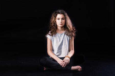A portrait of a cute girl teenager with long curly hair sitting in a lotus posture on dark background.の写真素材