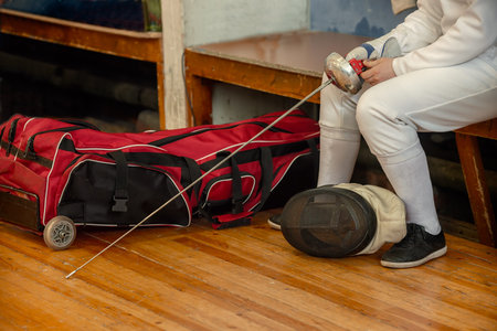 Girl participant in the fencing competition on swords sits on a bench holding a sword in her hand,...