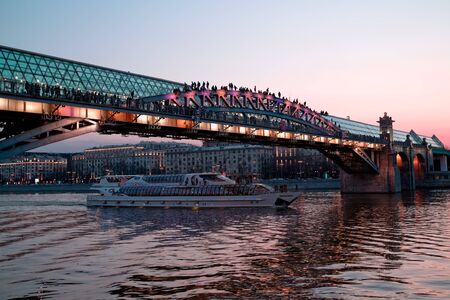Russia, Moscow, April 15, 2018. Tourist excursion ship passes under the bridge over Moscow river. On the transparent roof of the bridge, people sit in spring and summer evenings at sunsetのeditorial素材