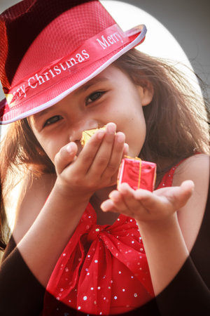 A girl in a red dress wearing a red hat symbol of christmas mischievous cheerful holding a small gift boxの写真素材