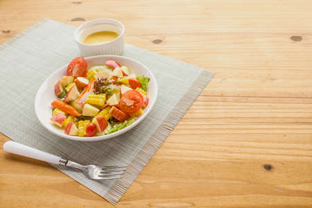 Mixed fruit and vegetable salad, crab sticks on a white plate, salad bowls on the wooden floor, leaving some empty space.の写真素材