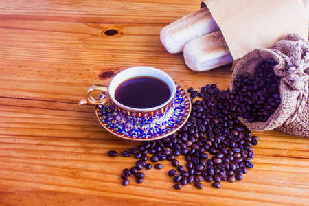 Black coffee in a beautiful patterned cup with a bag of coffee beans and a bag of bread.の写真素材