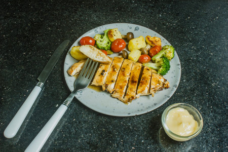 Grilled chicken breast, beef, steak, potatoes, cherry tomatoes, broccoli on a black plate, sauce cup and fork on black background.の写真素材