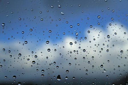 Raindrops on a window pane against a blue sky with cloudsの写真素材
