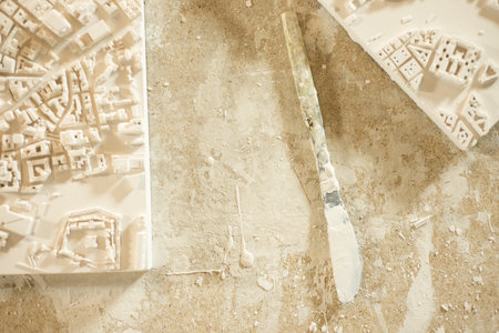 A plaster-coated knife placed between two 3D city map plaster models on plaster-splattered wooden...