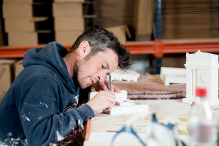 A man in paint-splattered dark hoodie busy engraving a section of a white plaster building model...