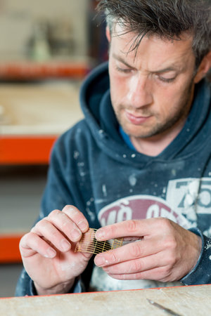 A stubbly caucasian male in paint-splattered carefully examining a part of product.