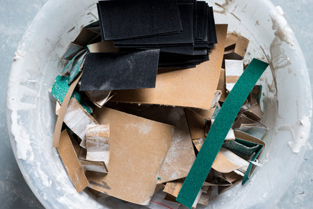 Closeup of strips of used and unused sandpaper in a plaster-laced plastic bowl on stone surface