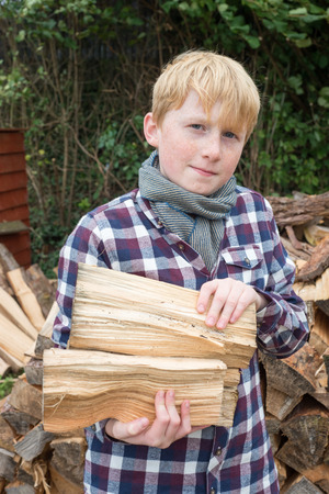 Boy in plaid shirt, scarf, and hat holding an axe in front of a pile of firewood in a farmの写真素材