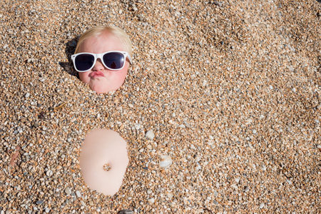 A young blonde girl in sunglasses pulls a funny face to camera from beneath the pebbles of a beach. Everything except her head and belly button has been buried beneath the stonesの写真素材