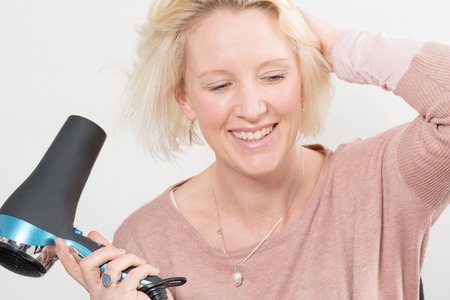Blonde lady smiling while drying her hair with an electric blow dryer over a white backgroundの写真素材