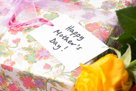 Gift tag that reads "Happy Mother's Day" placed on a wrapped present beside a yellow rose.の写真素材