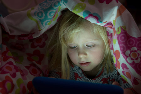 Little girl with a blanket over her head using a tablet in the dark