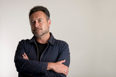 Bearded adult man in a blue jacket with arms crossed over a gray background with copy space