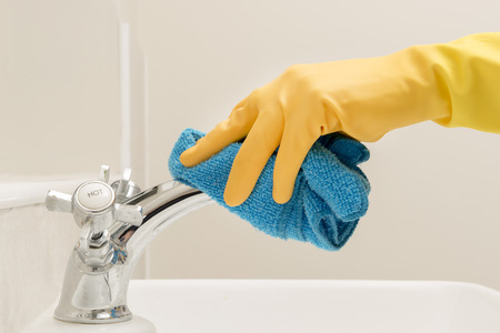Hand wearing a yellow rubber glove wiping a faucet with a blue rag in a washroomの写真素材