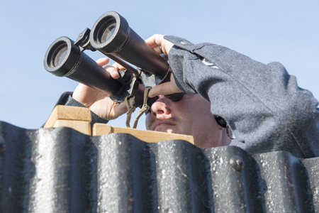Male military personnel peering through a pair of huge binoculars behind a corrugated iron sheetの写真素材