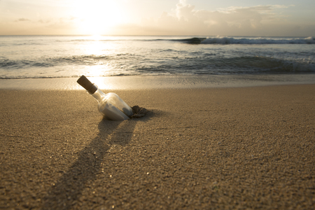 Message in a bottle half-buried in sand with ocean, sky and sunset in backgroundの写真素材