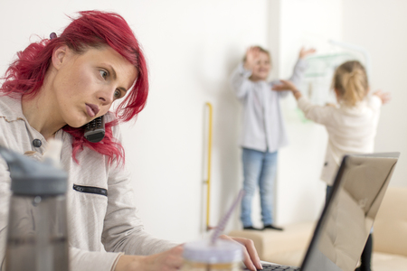 Red-haired mom on the phone while typing on a laptop as kids play in the backgroundの写真素材