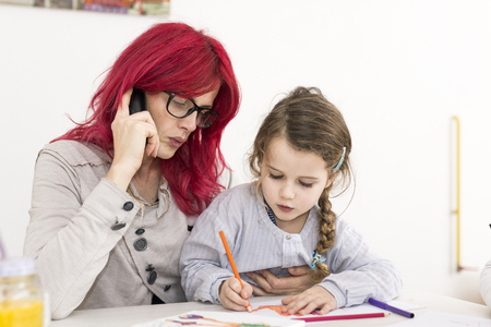Woman talking on the phone as her kid sits on her lap while drawingの写真素材