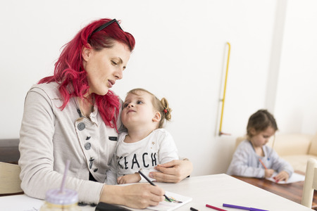 Woman drawing art with her on her lap as another kid doodles on a table in the backgroundの写真素材