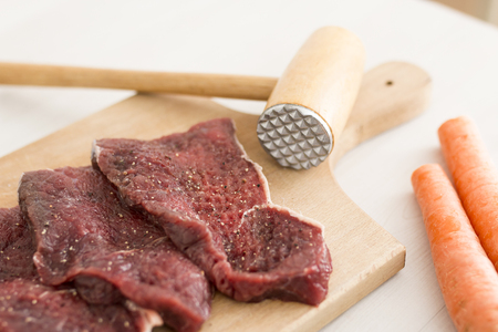 Close-up of seasoned raw meat slices on a wooden cutting board with carrots and a wooden meat tenderizerの写真素材