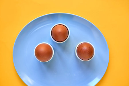 Three brown eggs in individual egg cups on a blue plate over a yellow background