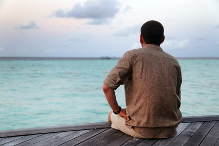 A lonely man is sitting on a wooden pier and looking into the ocean distance.の写真素材