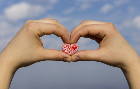 Two female hands are holding a bright red speckled ceramic heart against the blue sky with clouds.の写真素材