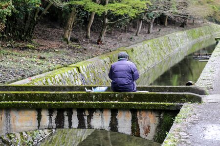 Fisherman preparing bait sitting on a bridge above a channelの写真素材