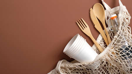 Zero waste concept. Reusable produce bag with wooden cutlery, glass bottle and carton cups on brown background. Eco-friendly, biodegradable, plastic free tableware. Sustainable lifestyle.の写真素材