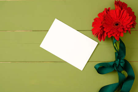Bouquet of red gerberas and blank paper card on pastel green wooden background. Flat lay composition, greeting card mockup with flowers. Flat lay, top view, overhead.の写真素材