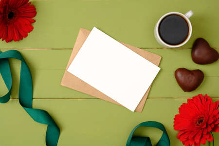 Blank paper card and envelope, coffee cup, flowers, ribbon on pastel green background. Feminine accessories on the wooden table. Creative layout, beauty blogger desk concept. Top view, overhead.の写真素材